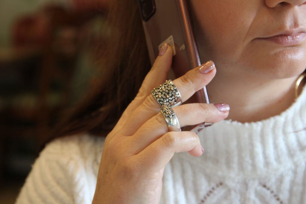 In this image is a woman with her mobile phone in her hand. She is sitting in a coffee shop and holding the phone to her ear which shows the rings on her hand. The rings are silver oxidised, with a bit of gold and daisy details.